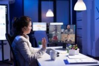 Back view of business woman talking about sale report in video conference working overtime in start-up office. Team using online wireless communication discussing on webcam during group meeting.