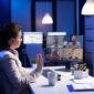 Back view of business woman talking about sale report in video conference working overtime in start-up office. Team using online wireless communication discussing on webcam during group meeting.