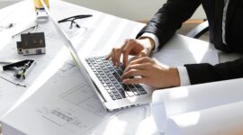 Hands of CEO of construction company in black suit typing message via e-mail to partners on laptop with drawings, seal stamp, divider on table. Young architect using netbook sitting in his office