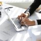 Hands of CEO of construction company in black suit typing message via e-mail to partners on laptop with drawings, seal stamp, divider on table. Young architect using netbook sitting in his office