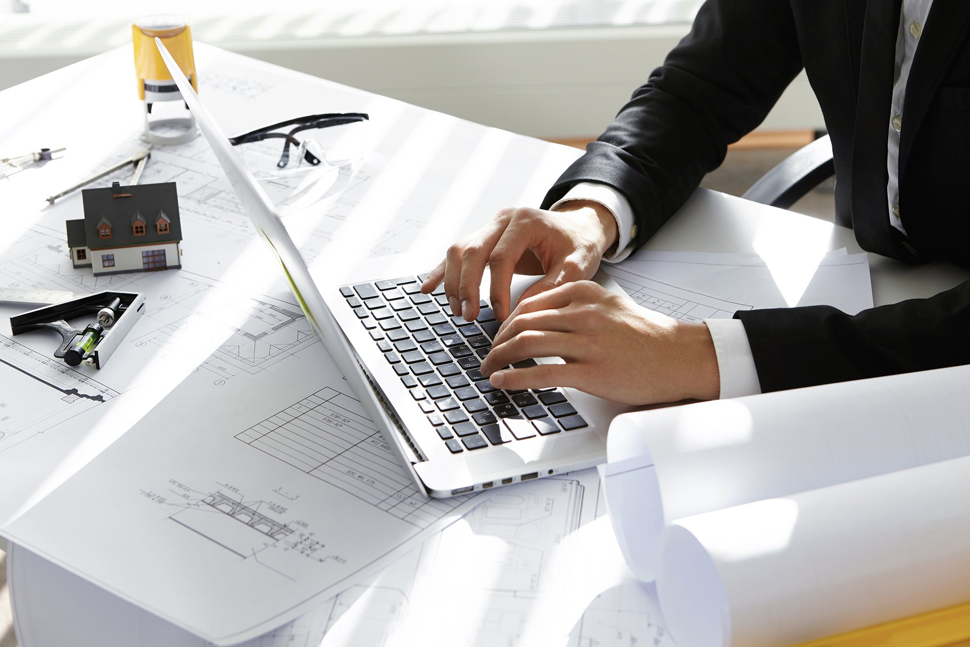 Hands of CEO of construction company in black suit typing message via e-mail to partners on laptop with drawings, seal stamp, divider on table. Young architect using netbook sitting in his office